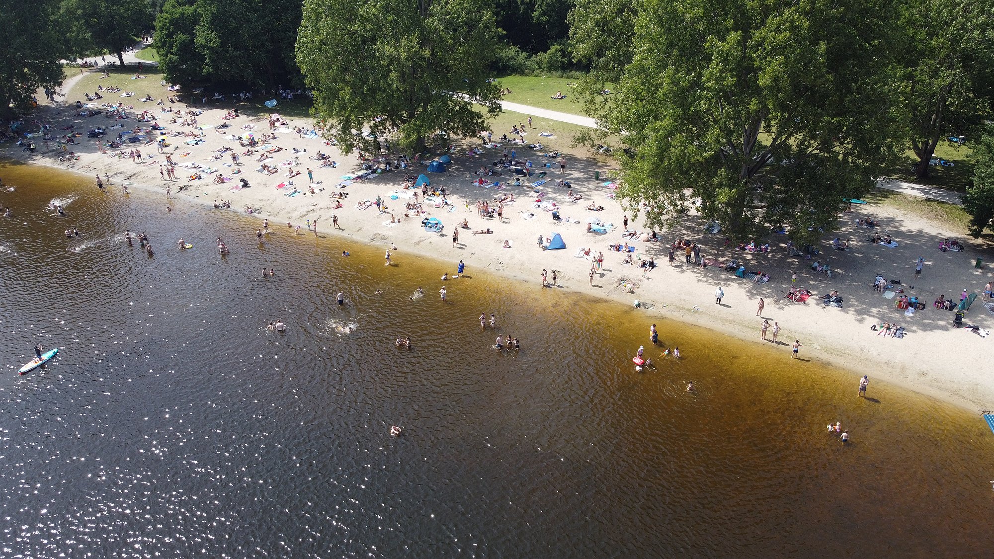 Bild beinhaltet eine Draufsicht auf den Altwarmbüchener See in Hannover. Es ist ein warmer Sommertag mit vielen Badegästen am Strand.