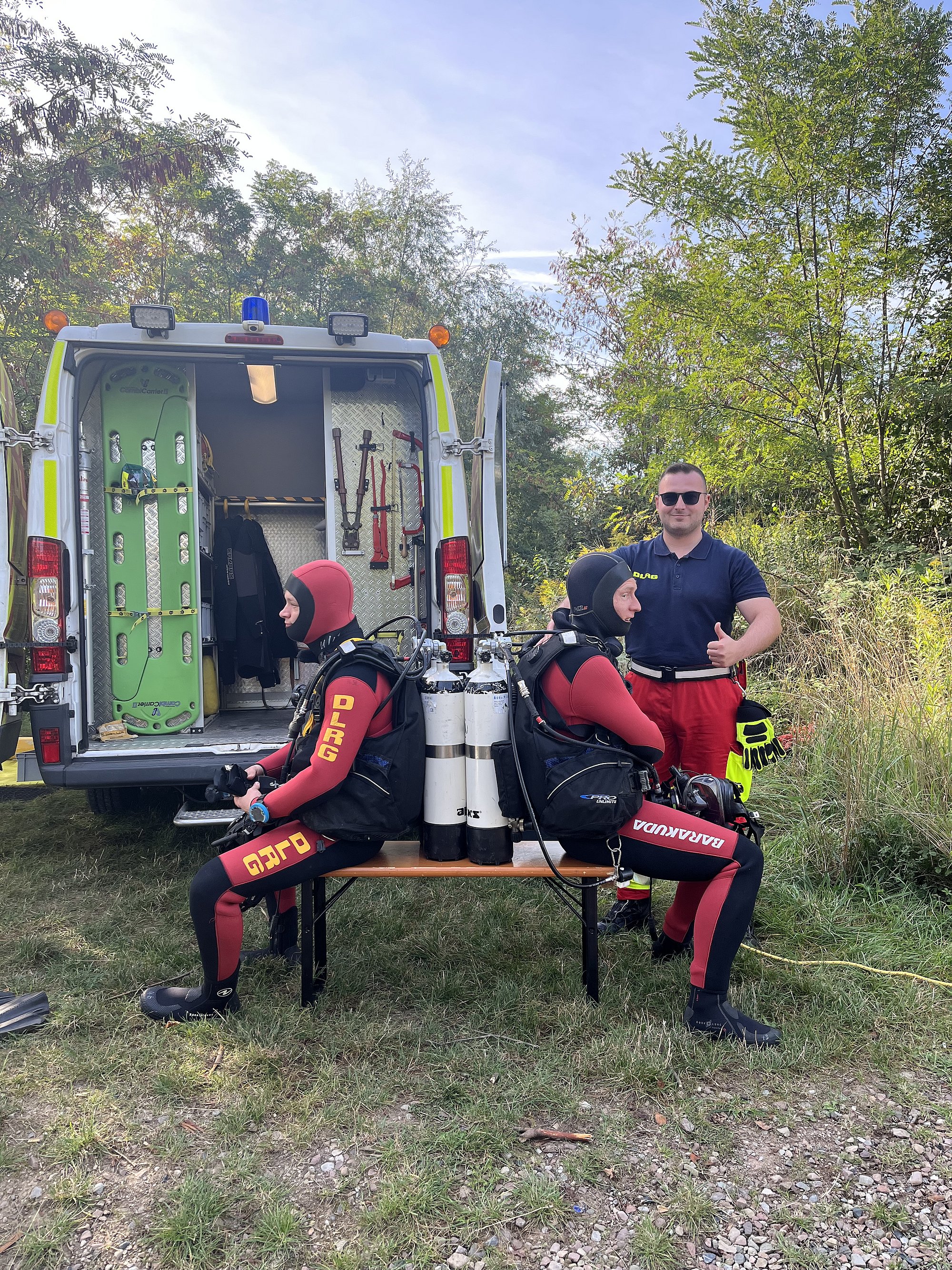 Bild beinhaltet zwei Taucher vor einem Auto sitzend auf einer Bank. Ein Signalmann steht gut gelaunt dahinter. Die Taucher sind vollständig ausgerüstet bereit für den Einsatz.