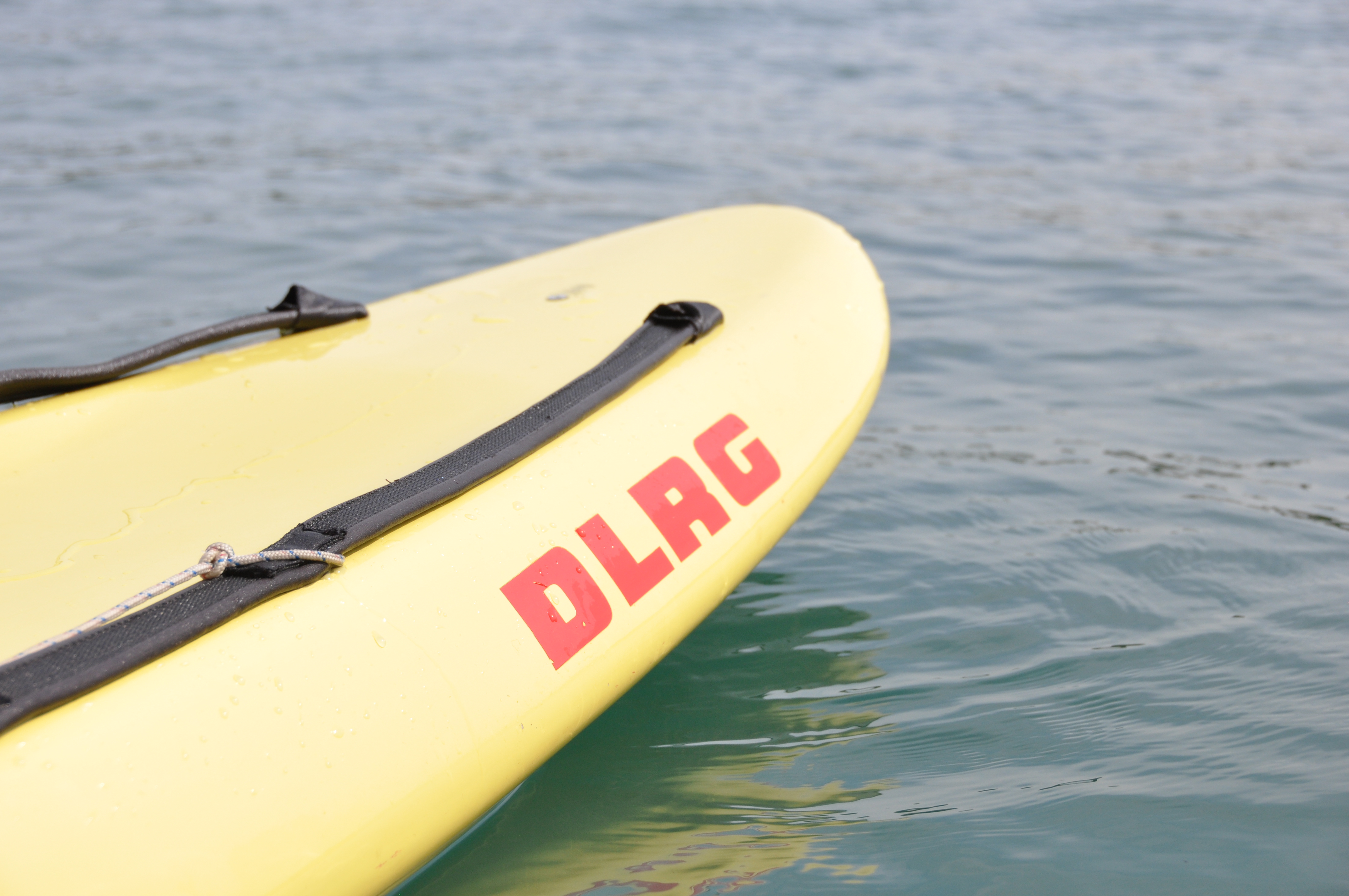 Bild beinhaltet ein Rettungsbrett in gelb, welches auf der Seite den DLRG Schriftzug stehen hat. Dieses Brett befindet sich auf dem Wasser.