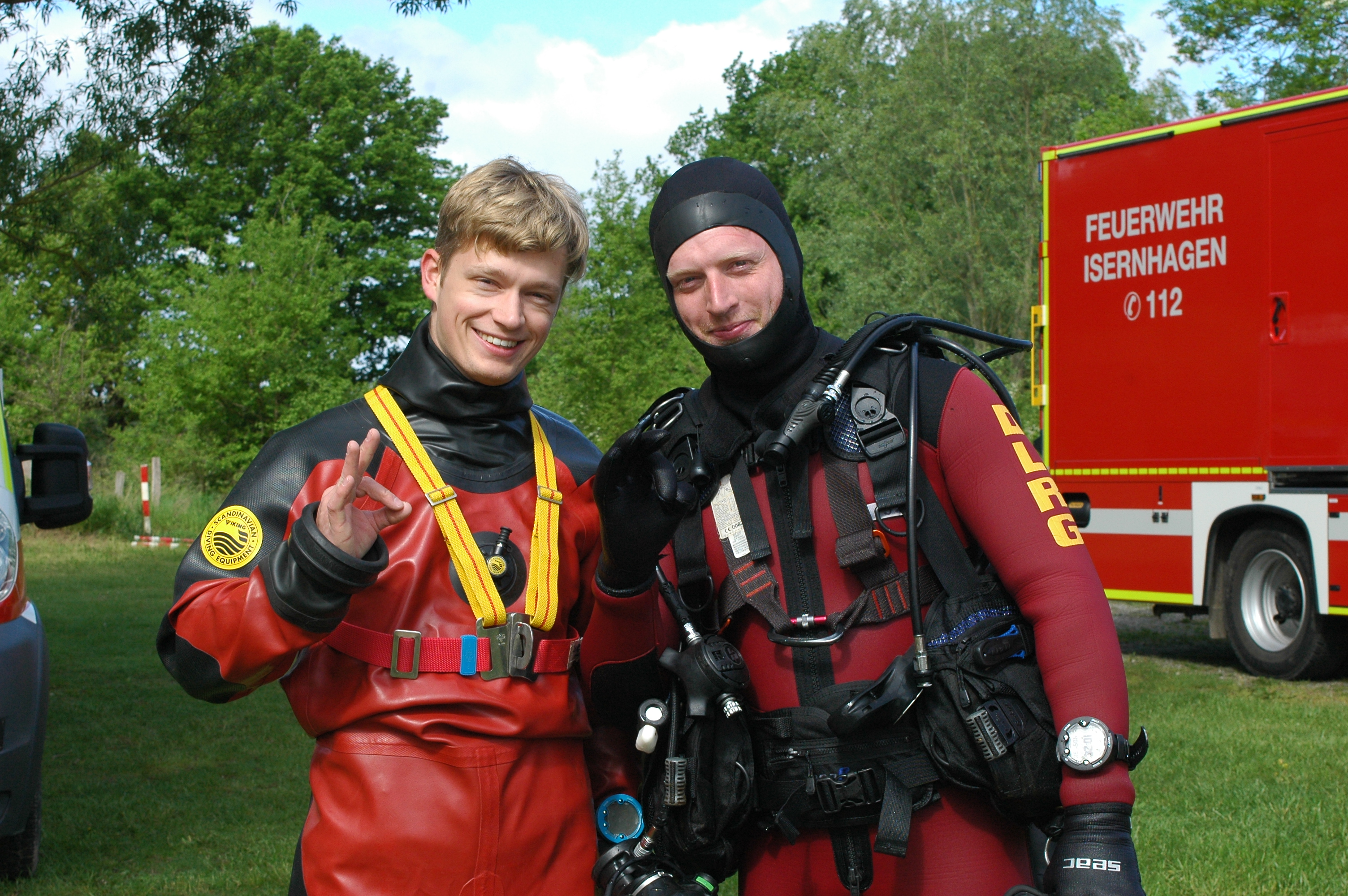 Bild beinhaltet zwei junge motivierte Taucher. Links ein Taucher mit einem Trockentauchanzug, der in jeder Art von Wasser tauchen kann, ohne dass er in Berührung damit kommt - deswegen Trockentauchanzug. Rechts steht ein Taucher im Nassanzug, welcher die komplette Ausrüstung trägt und auch freundlich in die Kamera blickt.