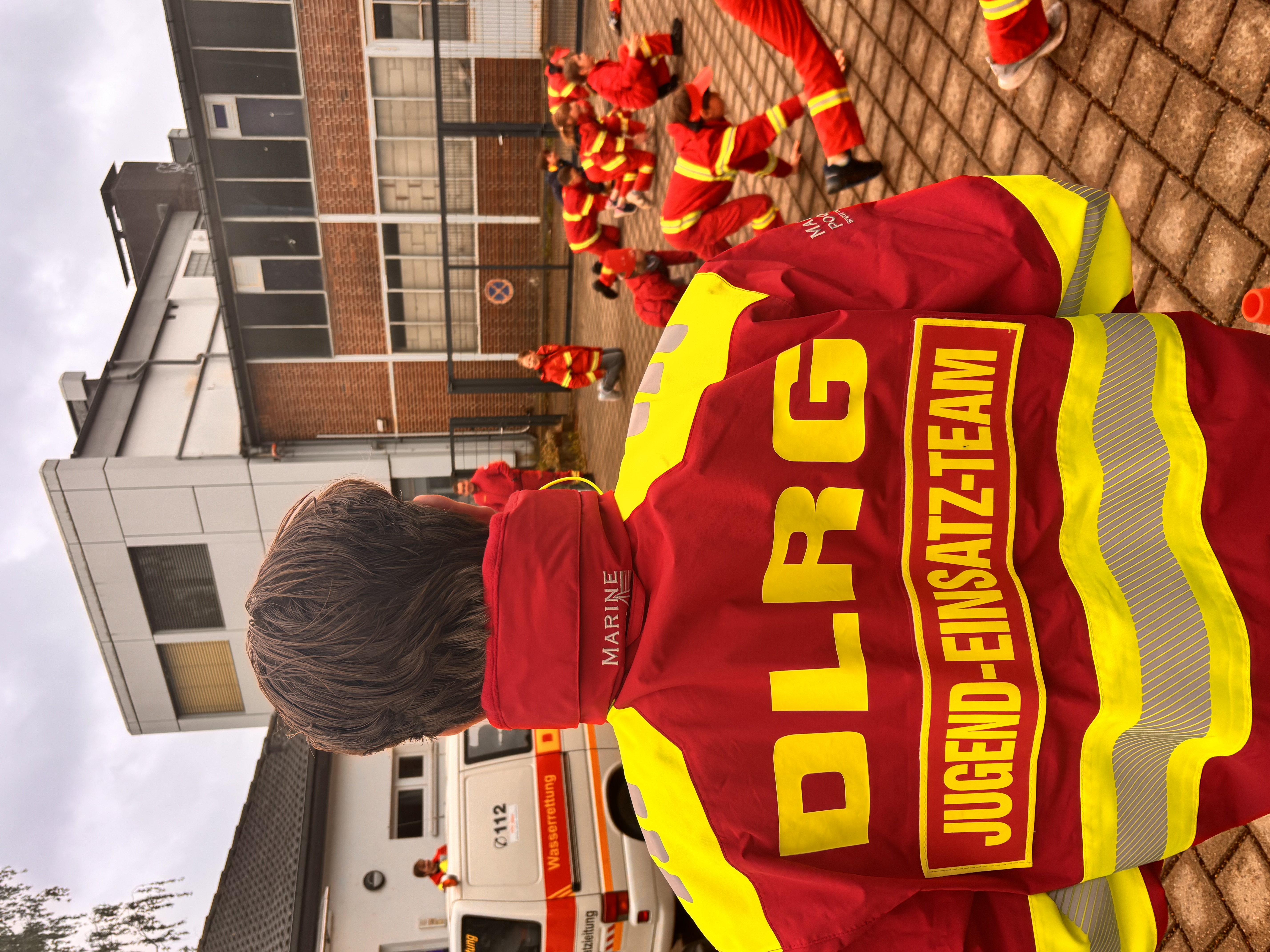 Bild beinhaltet einen Jugendlichen, der die DLRG-Einsatzkleidung trägt und auf seinem großen Rückenschild Jugend-Einsatz-Team stehen hat. Im Hintergrund spielen mehrere Jugendliche und Kinder ein Spiel mit Ganzkörpereinsatz. 